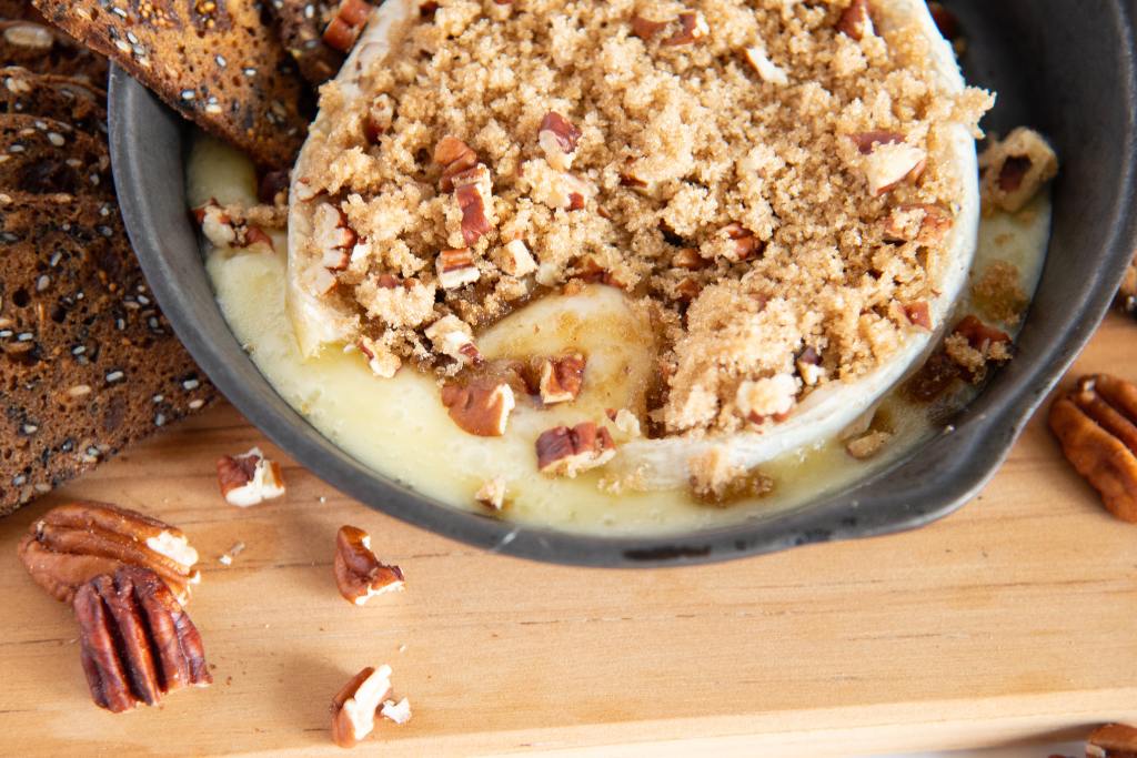 baked brie with brown sugar and chopped pecans on top, the brie has been cut open, so you can see the melty cheese inside it.