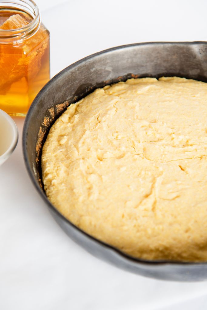 uncut cornbread in a cast iron skillet, you can see part of jar of honey next to it