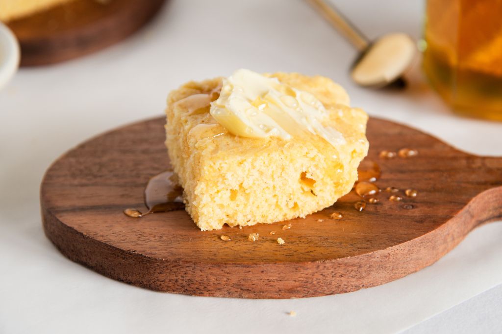 a close up of a piece of cornbread on a little wooden dish, there is butter and honey on top of the bread.