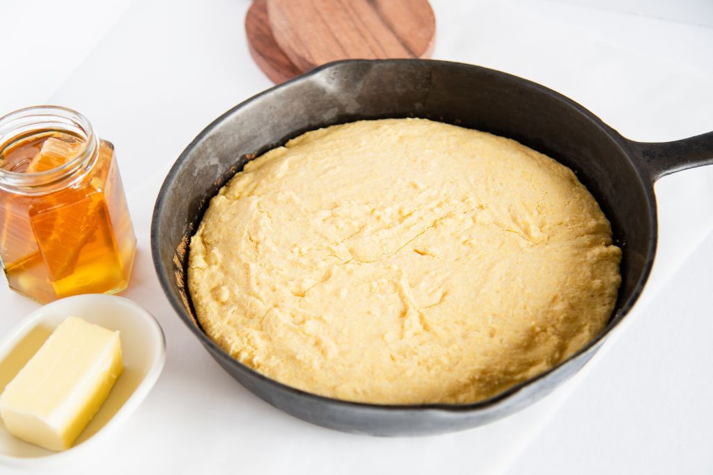 uncut cornbread in a cast iron skillet, there is a dish of butter and a jar of honey next to it