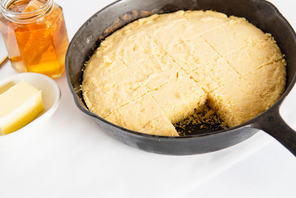 cut cornbread in a cast iron skillet, one piece has been taken out. There is a small dish of butter and a jar of honey next to it