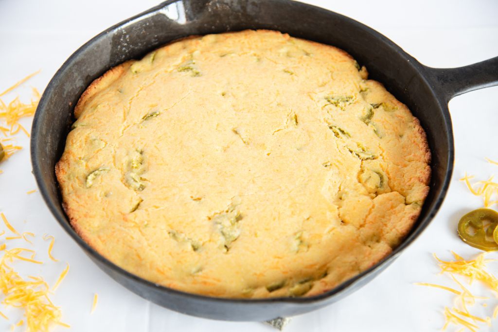 uncut jalapeno cheddar cornbread in a cast iron skillet, there is some shredded cheese and some sliced jalapenos around the skillet