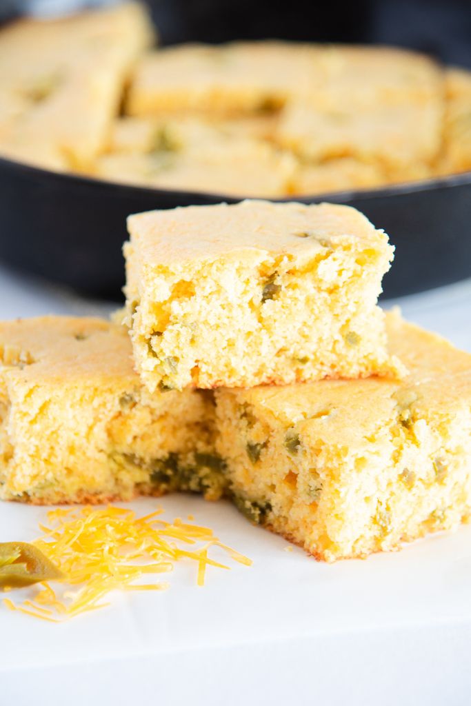 a piece of cornbread set on top of two other pieces, there is some shredded cheese in front of it, and you can see a skillet of cornbread behind it