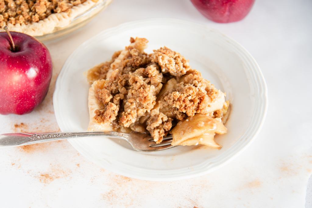 a slice of dutch apple pie on a plate with a fork on it. You can see a couple whole apples, and the rest of the pie in the background 