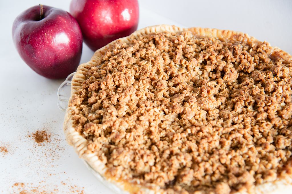 an uncut dutch apple pie, there are two whole apples next to it and some ground cinnamon sprinkled around it