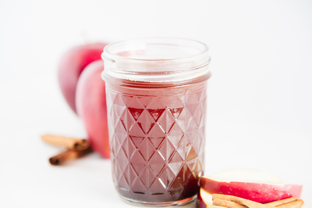 a jar of apple syrup with apple slices and cinnamon sticks next to it, there are a couple apples behind it too.