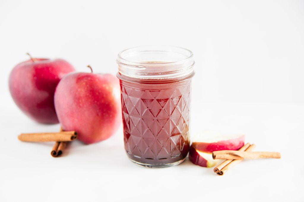 a jar of apple syrup,with apples, apple slices and cinnamon sticks next to it