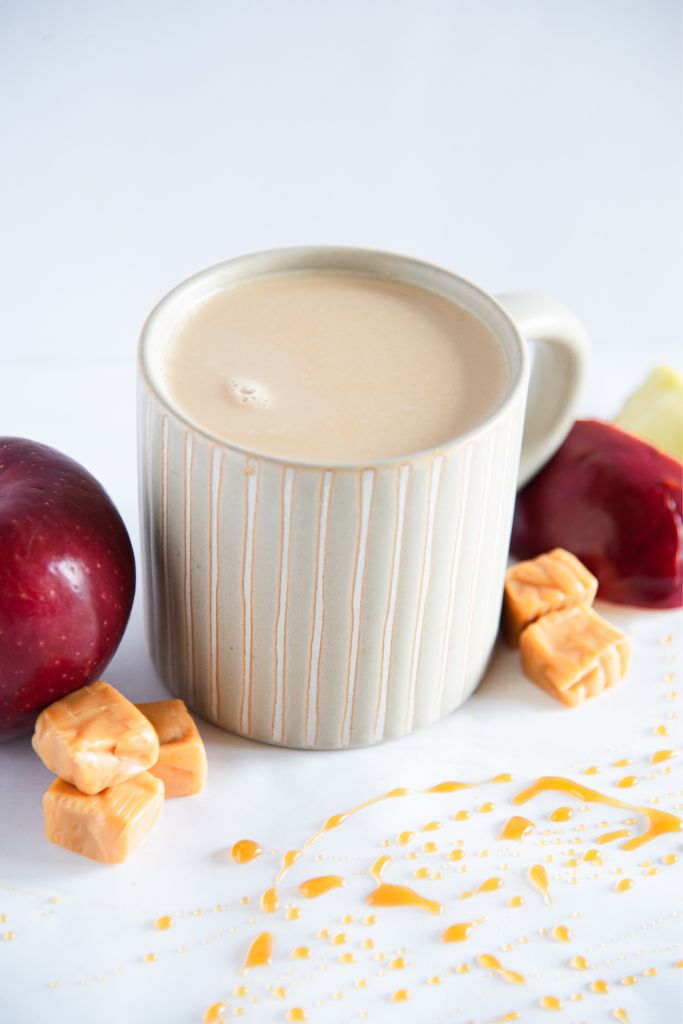 a finished latte with apples and caramel pieces around it, there is also some caramel drizzle in front of it