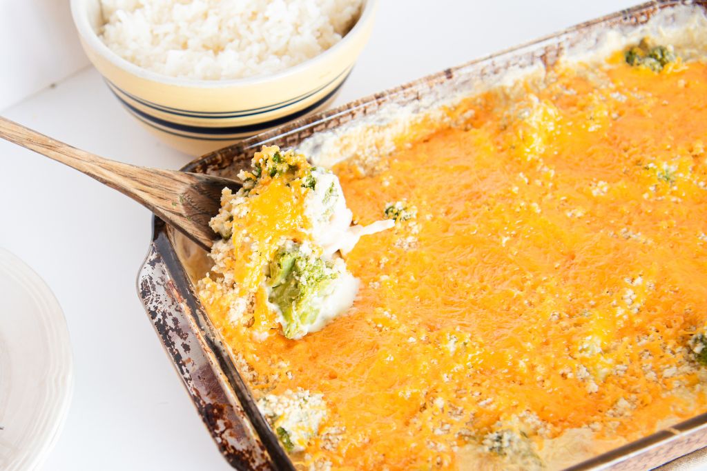 a scoop of chicken divan being taken out of the pan with a wooden spoon, there is a bowl of rice next to the pan