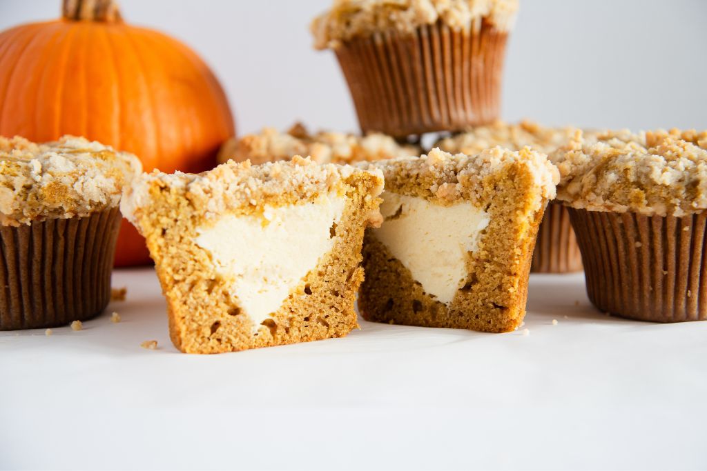 a muffin cut in half so you can see the cheesecake filling, there some whole muffins and a pumpkin behind it