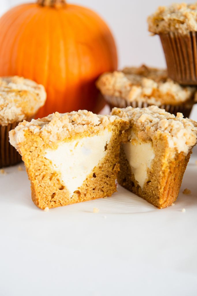 a pumpkin cheesecake muffin that has been cut in half, there some muffins and a pumpkin behind it