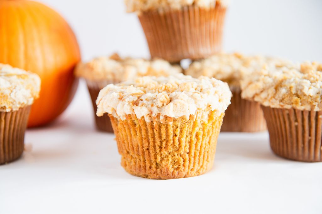 a close up of a pumpkin muffin, there are three muffins stacked up behind it, and a pumpkin to the left of it