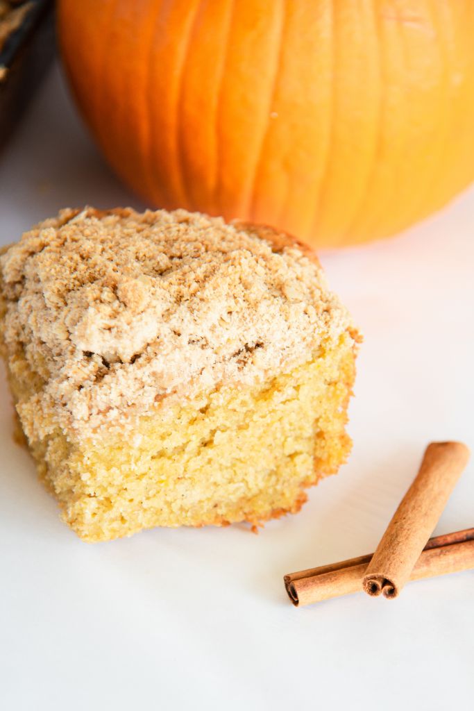 a close up of a piece of coffee cake with cinnamon sticks next to it, you can see part of a pumpkin behind it