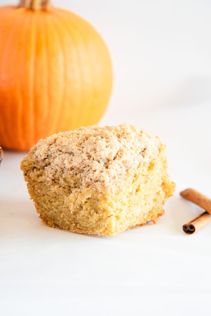 a piece of coffee cake with two cinnamon sticks to the side of it, there also a pumpkin behind it