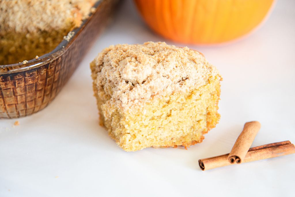 a piece of coffee cake with two cinnamon sticks next to it. You can see part of a pumpkin and a pan of coffee cake behind it