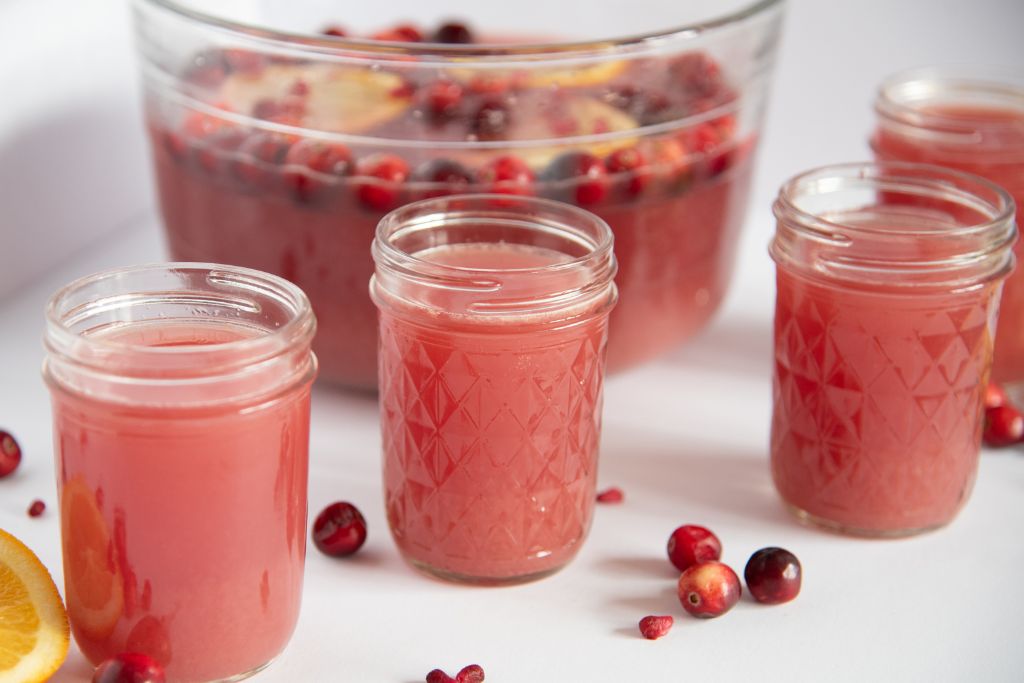 four glasses of punch in front of a bowl of punch, there are cranberries and pomegranate seeds around them 