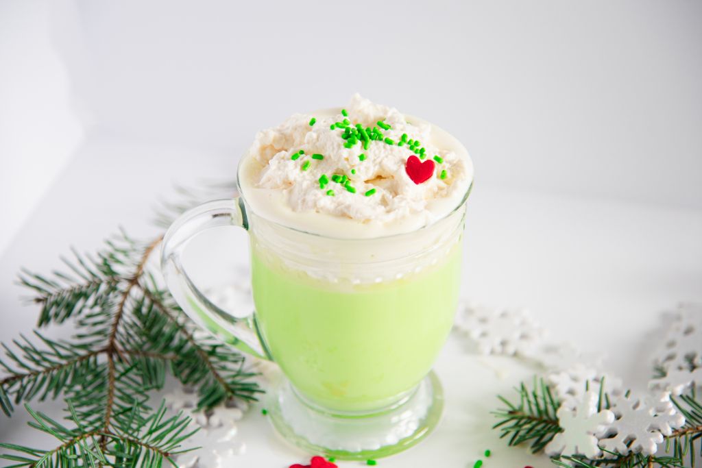 a cup of Grinch hot chocolate with pine branches and snowflakes around it