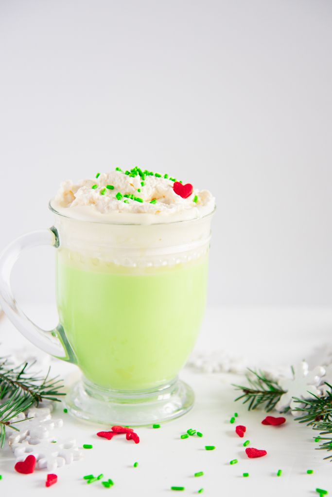 a cup of Grinch hot chocolate with pine branches, snowflakes, and sprinkles around it