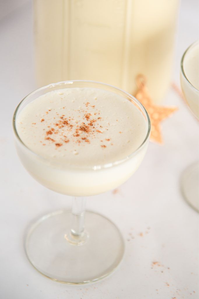 a glass of eggnog with ground nutmeg on top, there is a jar of eggnog and a gold star behind it 