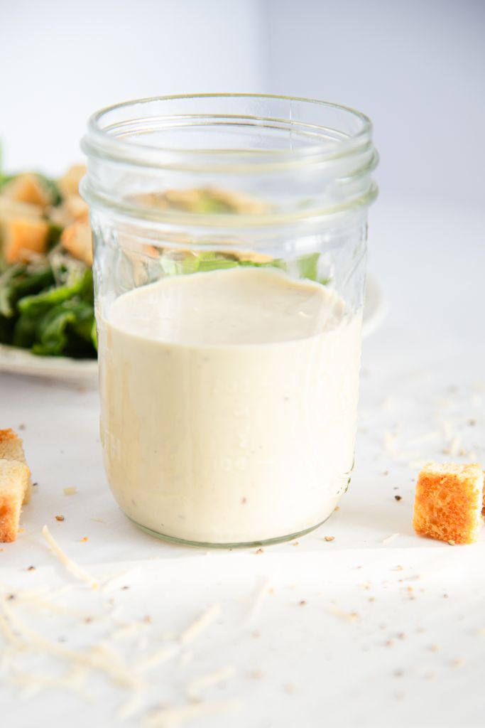 a jar of Caesar salad dressing with a plate of salad behind it