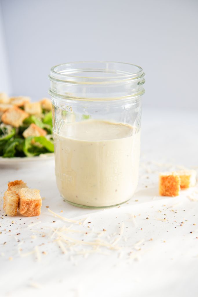 a jar of Caesar dressing with a plate of salad behind it, there some croutons around the jar too 