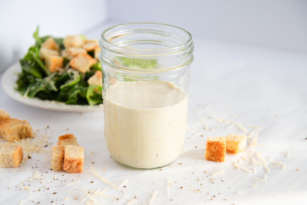 a jar of Caesar salad dressing with a salad behind it, there is some cheese and croutons around the jar too