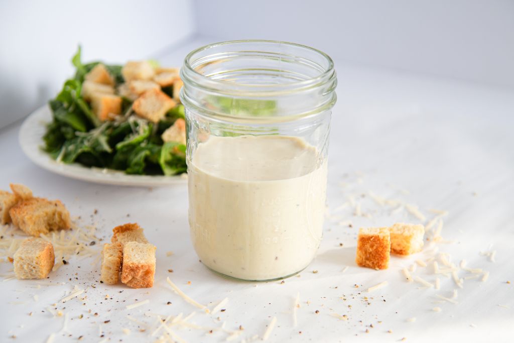 a jar of Caesar dressing with cheese and croutons around it, there is a plate of salad behind the jar