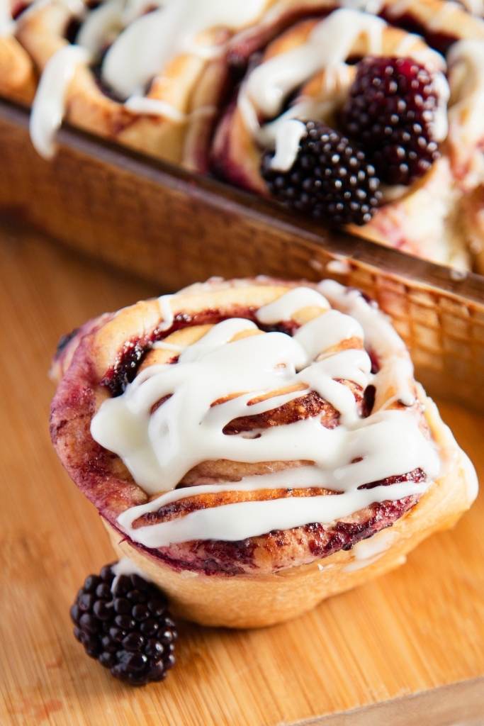 a close up of a sweet roll on a wooden dish, you can see a pan of sweet rolls behind it
