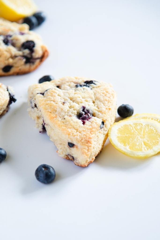 a blueberry lemon scone with lemon slices and blueberries next to it, you can also see more scones behind it 