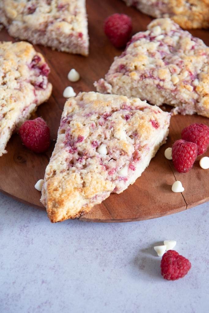 a close up of a white chocolate raspberry scone on a wooden plate, you can see more scones behind it