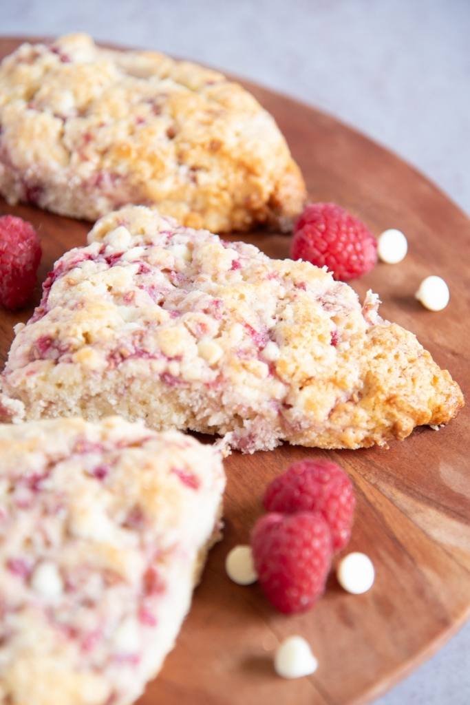 a close up on a scone on a wooden plate, there are more scones, raspberries and chocolate chips by it 