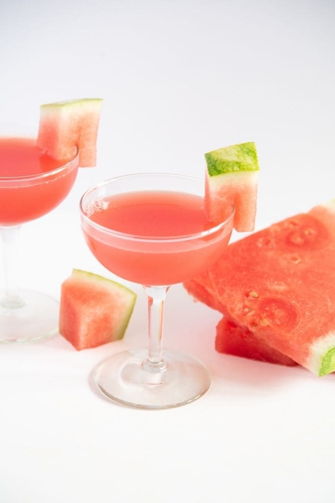 a finished cocktail with another cocktail behind it and watermelon slices next to it 
