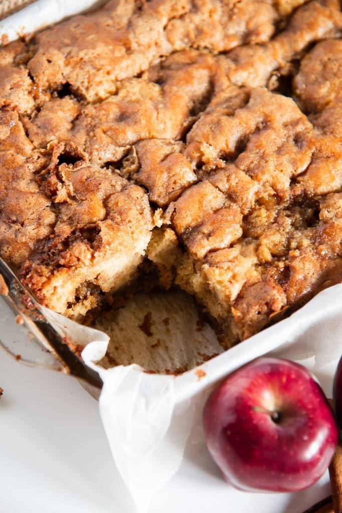 a pan of coffee cake with a piece taken out it, there is an apple next to the pan as well