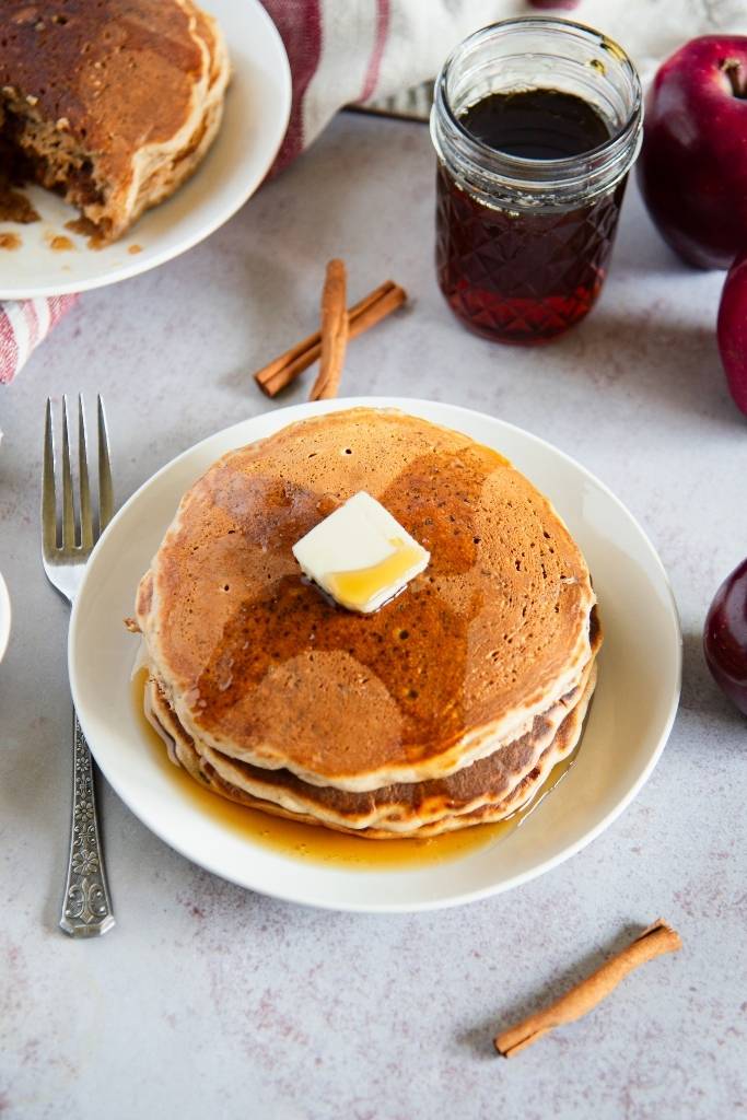 a plate of pancakes with butter and syrup on top, there are apples, cinnamon sticks and syrup around the plate 