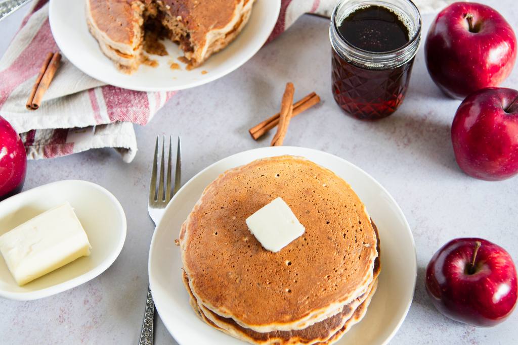 a plate of pancakes with butter on top, there is syrup, butter, apples, and more pancakes around the plate 