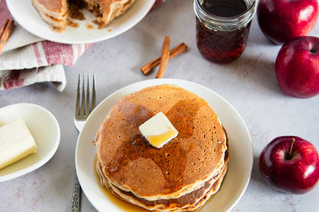 a stack of pancakes with butter and syrup on top, there are apples, more pancakes, a jar of syrup and a dish of butter next to the plate of pancakes 