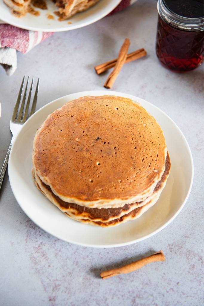 a plate of pancakes, there is a fork and some cinnamon sticks next to the plate, you can also see a jar of syrup behind the plate 