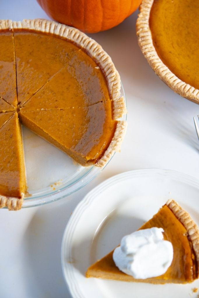 a pumpkin pie with a slice taken out of it, next to a plate with a piece of pie with whipped cream on top