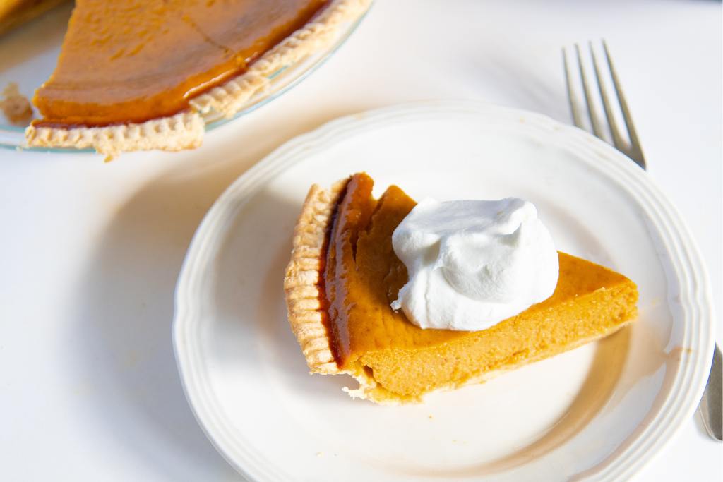 a slice of pumpkin pie with whipped cream on a plate, you an see part of pumpkin pie behind it