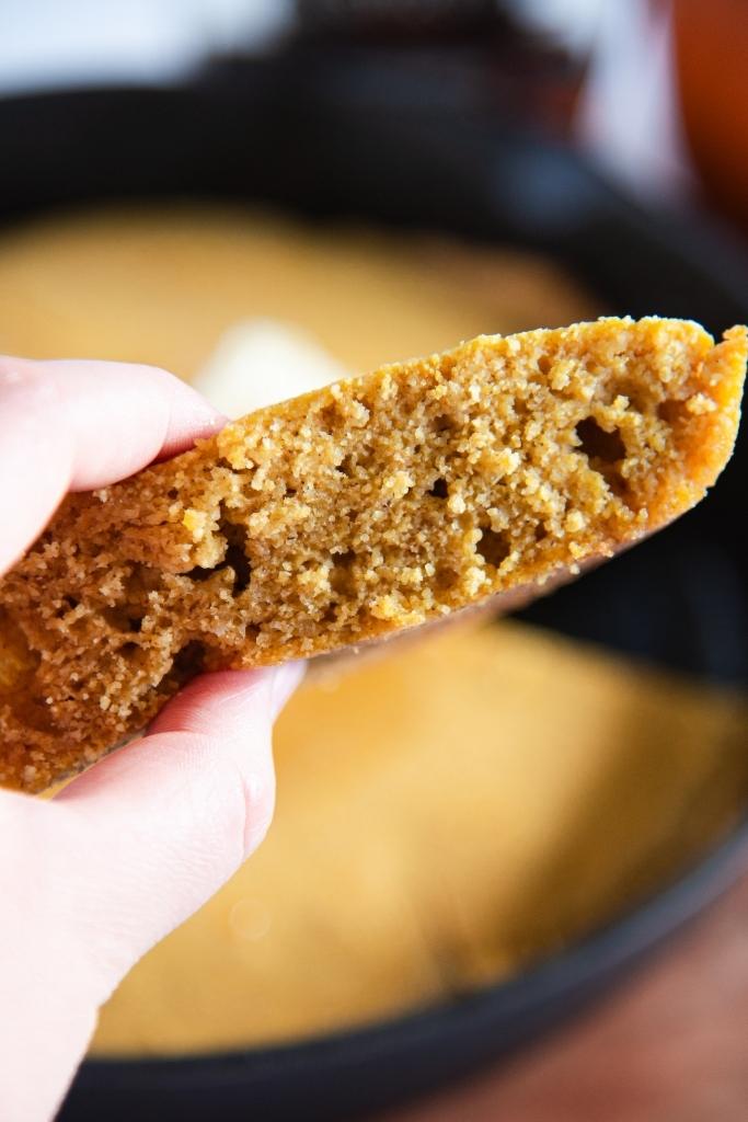 a close up of a slice of cornbread bread being held in front of the skillet of cornbread 