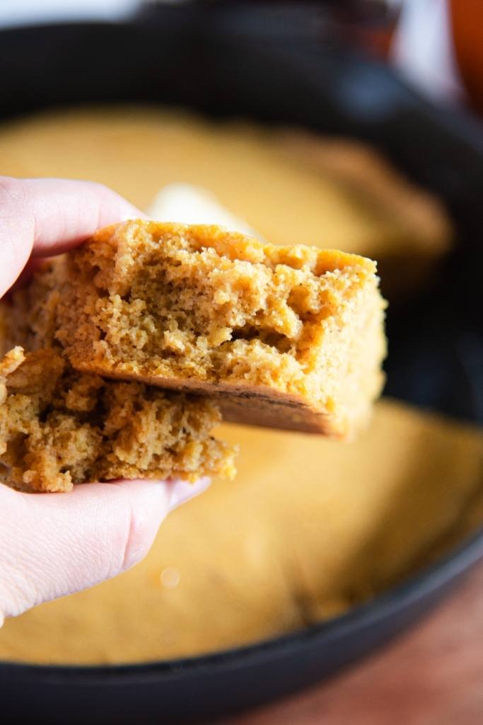 a broken in half piece of cornbread being held in front of a skillet of cornbread 