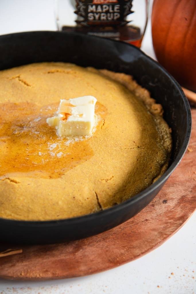 a skillet of uncut cornbread with butter and maple syrup on top. You can see part of a pumpkin and a bottle of syrup behind it 