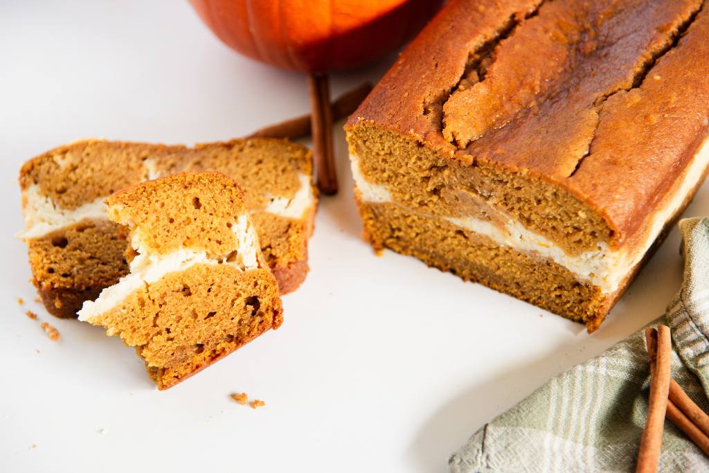 a loaf of pumpkin bread that has been cut off of, there are some slices of bread and cinnamon sticks next to it 