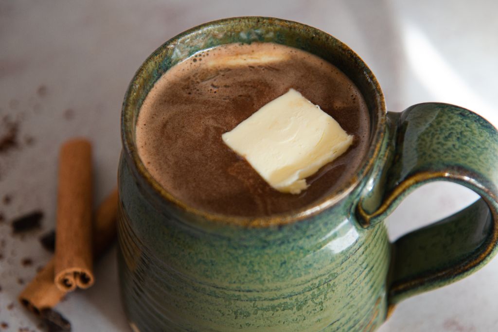 a close up of a cup of buttered rum hot chocolate, with a pat of butter on top and cinnamon sticks next to it 