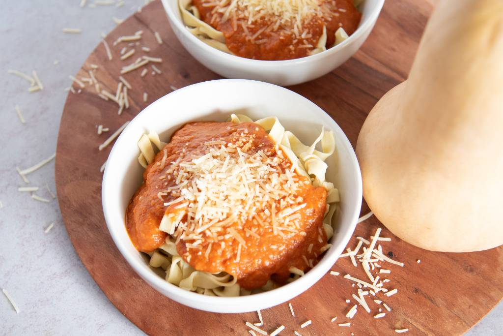 a bowl of noodles with sauce on top, there is a whole squash next to the bowl and a bowl of noodles behind it 