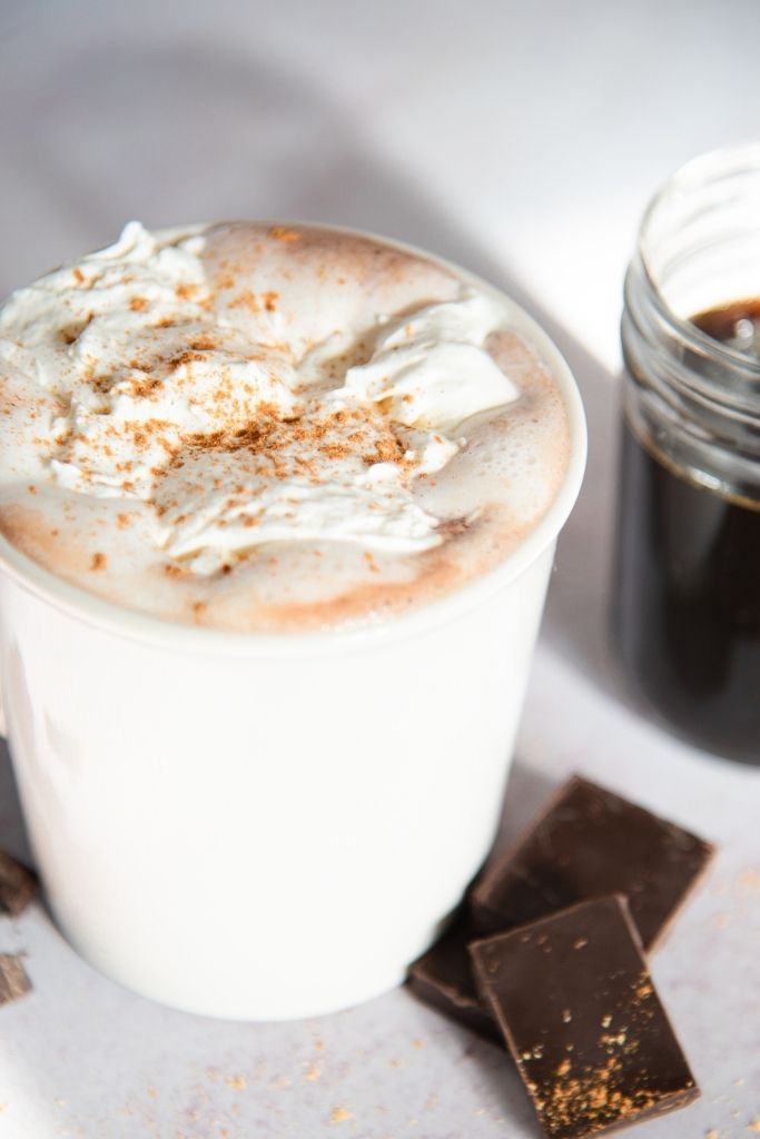 a cup of hot chocolate with pieces of chocolate next to it. There is also a jar of gingerbread syrup next to the cup