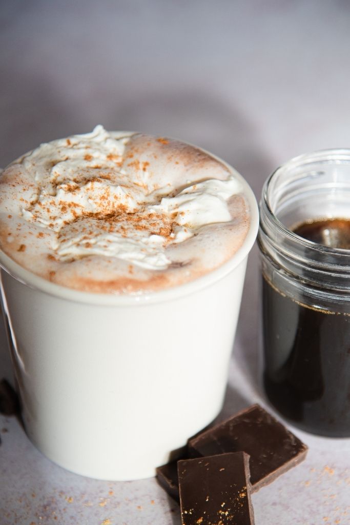 a close up of a cup of hot chocolate with a jar of syrup and some pieces of chocolate next to it 