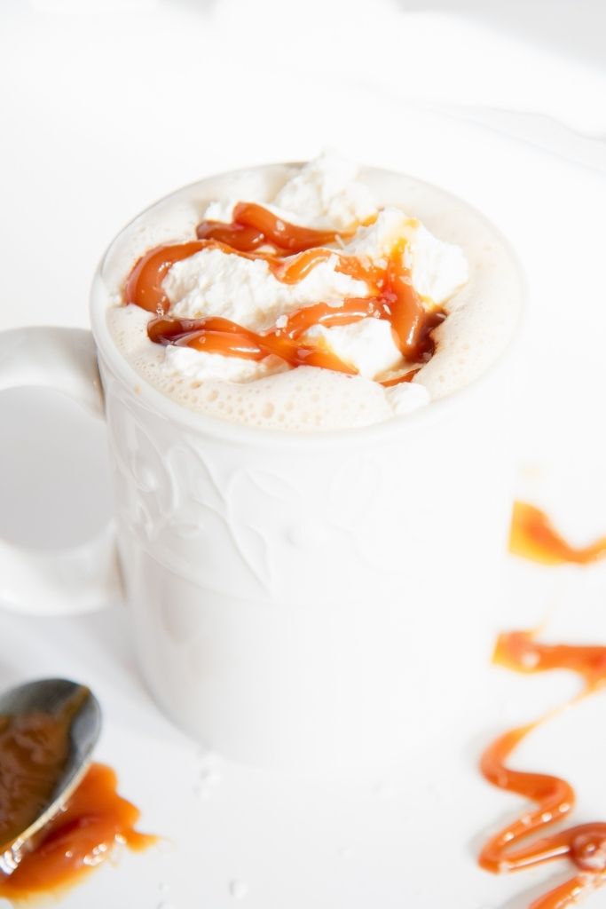 a close up of a finished latte, there is a spoonful of caramel and a caramel drizzle next to the cup