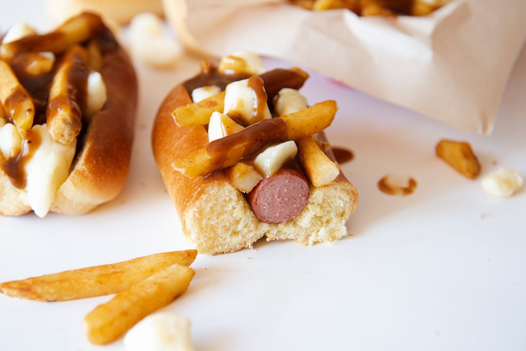 a poutine topped hot dog that has been cut in half, there are some french fries and cheese curds next to it 
