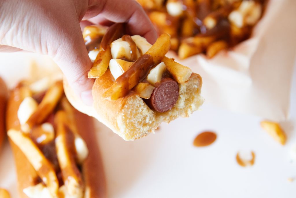 someone holding a half of a hot dog, you can see another topped hot dog, and plate of fries behind it 